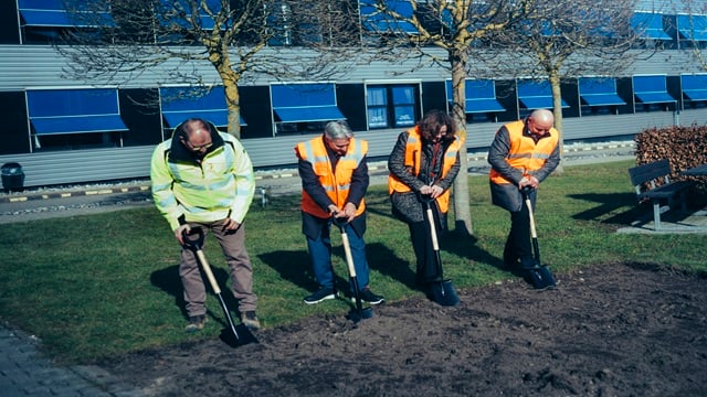 Four people in safety vests digging with shovels during a ceremonial groundbreaking in front of a building