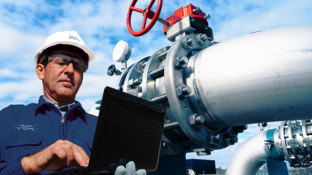 engineer wearing hard hat and safety glasses holding laptop in front of pipes and blue sky